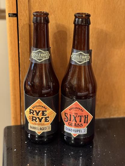 Two brown bottles of beer. To the left a light orange diamond shaped label with dark brown lettering and tan accents. To the right a dark orange label with light blue banner. Both have Boulevard neck labels