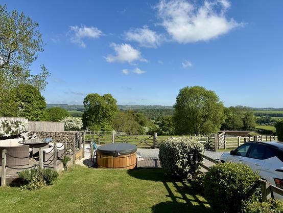Under a bright blue sky dotted with soft white clouds, the rolling countryside of Leyburn stretches across the horizon in a vibrant patchwork of greens. From a neatly kept garden at Highfield House, a cozy outdoor seating area and a round wooden hot tub overlook open fields bordered by wooden fences and hedgerows. A white car rests in the gravel drive nearby, while lush trees and distant hills frame the serene rural view. Sunlight casts clear shadows across the trimmed lawn and garden shrubs, adding warmth to the peaceful, open landscape that extends toward the horizon.