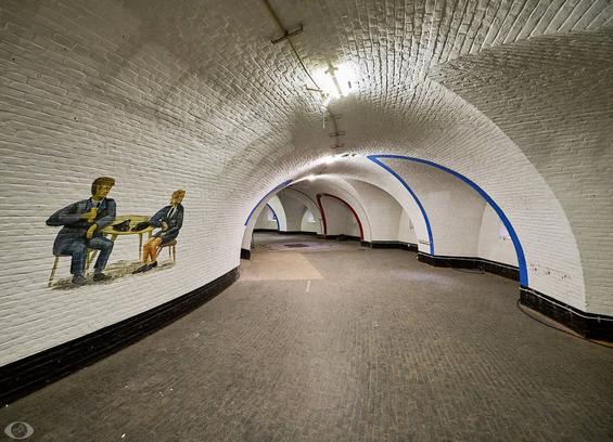 A corridor in a semicircle. The massive brick walls are painted white with a black border on the bottom. The left wall has a wall painting of a (presumably police-)uniformed man and woman sitting on a round table with the man apparently telling a story elbowed on the table and holding a glass in his right hand with the woman listening also having her richt elbow on the table. Their hats lie both on the table. To the right the wall has alcoves showing the thickness of the wall (80cm of solid brickwork.)