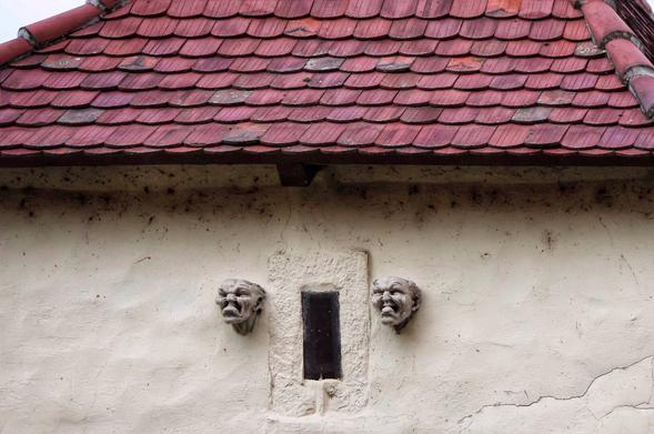A detailed, eye-level shot of a weathered, beige-coloured wall of an old building, featuring a small, rectangular window with a dark pane and a thick, stone frame. On either side of the window, two grotesque, sculpted stone heads with prominent wrinkles and exaggerated, grimacing expressions jut out from the wall. The head on the left has a more sinister, scowling look, while the one on the right appears to be snarling or laughing. Above, a red-tiled roof with a scalloped pattern is partially visible. The wall shows signs of age, with several fine cracks and some discolouration.