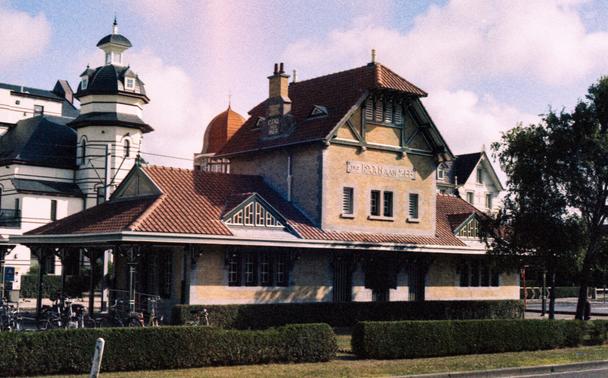 Color photo of the Kusttram station at de Haan, a building with a red-tiled roof and decorative gables, with the words “LE COQ SUR MER” and “DE HAAN AAN ZEE” displayed on its upper walls. Several bicycles are parked in front, and larger multi-story buildings with towers and varied rooftops stand in the background.
