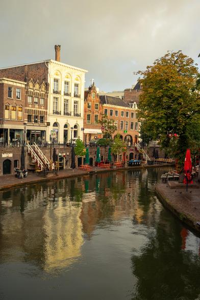 The image depicts a scenic view of a canal in a city, likely in Europe. Prominent buildings line one side of the canal, showcasing a mix of architectural styles. The structures range from ornate, historical designs with brick facades and decorative windows to more modern, streamlined forms. Many of the buildings have large windows and some feature balconies with railings.

Along the canal's edge on the left, there’s a row of outdoor seating areas equipped with tables and chairs, suggesting the presence of cafes or restaurants. The seating area is adorned with tables covered by umbrellas, including a conspicuous red one. Trees with green and some yellow leaves line the right side, adding a touch of autumnal color to the scene.

Reflections of the buildings and trees dance on the calm water of the canal, creating a serene and picturesque ambiance. The sky is lightly overcast with subdued lighting, and the overall atmosphere is tranquil, likely capturing a moment during the early morning. The image conveys a peaceful urban setting with a blend of natural and architectural beauty.