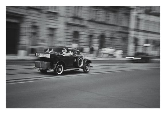 A black and white, horizontal, low-angle shot shows a vintage car driving from left to right on a street. The car is an open-top phaeton model with spoked wheels. The motion blur in the background suggests it's moving at a high speed. The car has a driver and two passengers, and a trunk is strapped to the back. A tram track runs parallel to the car's path, and buildings line the street.