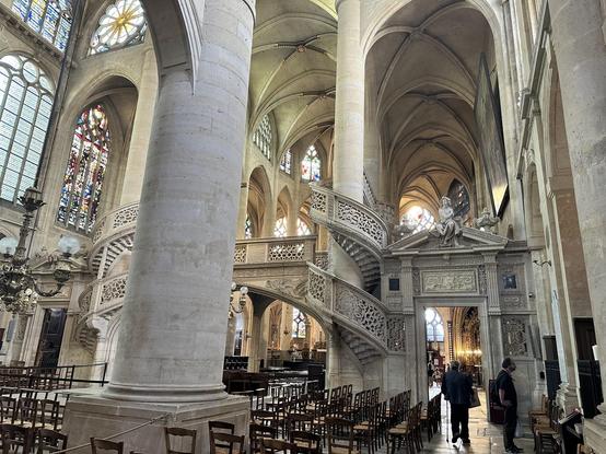 Oblique view across the nave, with parts of the columns, stained glass windows, ribbed vaulted ceiling and rows of chairs  visible. Visible in mid range is the elegantly-carved stone rood screen with equally ornate spiral staircases leading up to it.