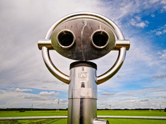 A telescope stands in front of a vast landscape with green meadows and a cloudy but blue sky. However, it is not long and thin, but rather a silver installation reminiscent of a Cyberman from Doctor Who. Someone has also painted eyelashes over the peepholes. It looks as if the telescope is looking at me.