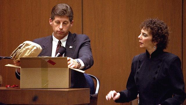Los Angeles police detective Mark Furhman being examined by prosecutor Marcia Clark during the O. J. Simpson trial, 10 March 1995. Fuhrman was later convicted of perjury for his testimony. Photo of a man in a suit in a witness stand examining the contents of a box; a woman next to him is speaking to him