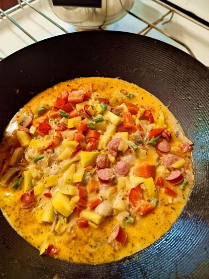 A wok with various chopped pieces of food simmering in it. The bits of food are yellow zucchini, red paprika, green beans and a darker yellow sauce. There's some bits of sausage and mushrooms swimming around, too.
