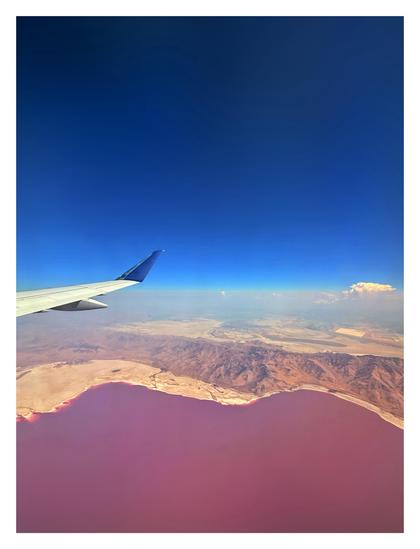 View from the window of an airplane flying over a vast lake with deep red water, bordered by a brown-hued mountain range. Above is a clear blue sky. The plane’s wing is at left frame. At right, a distant cloud floats over a layer of light haze.