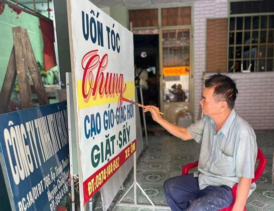 Man sat on a chair painting a large freestanding sign with Vietnamese lettering. Other signs are propped up against the wall behind this in what is a light industrial workshop setting.