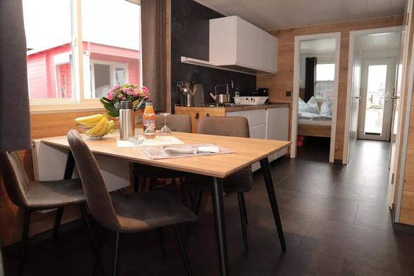 Alles klar – hier eine reine, sachliche Bildbeschreibung:

Die Bilder zeigen das Innere eines modernen Bungalowboots. Zu sehen sind ein Essbereich mit Holztisch, vier Stühlen, frischem Obst und Getränken, sowie eine Küchenzeile mit weißen Schränken, Spüle, Gasherd und Küchenutensilien. Dahinter liegt ein Schlafzimmer mit Doppelbett, grauen Vorhängen und Bettwäsche im Punktmuster. Der Grundriss zeigt zwei Schlafzimmer, ein zentrales Bad, die Küche mit Essbereich sowie Sitzgelegenheiten und Terrassen an Bug und Heck.