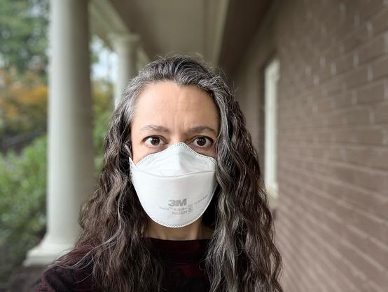 Middle-aged woman with long hair and N95 respirator outside on a columned walkway with trees in the background.