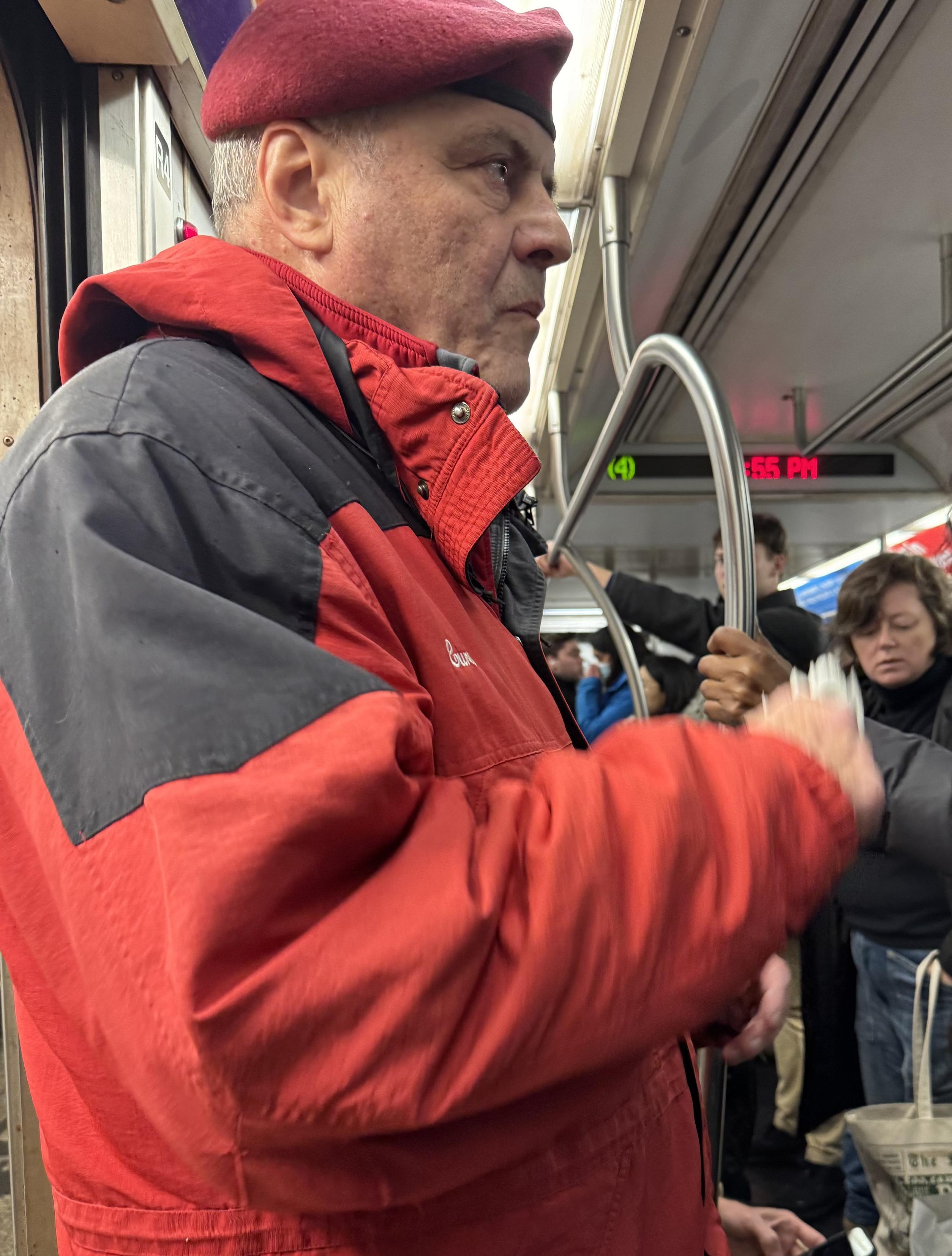 Failed NYC mayoral candidate and aspiring vigilante leader Curtis Sliwa in a subway car, dressed as in a red suit, vaguely resembling a petty, angry, clean-shaven Santa.