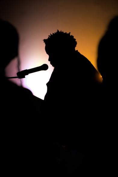 A backlit photo with the subject in shadow. The lighting is warm and the subject is sitting on a couch with a microphone, reciting Black revolutionary poetry