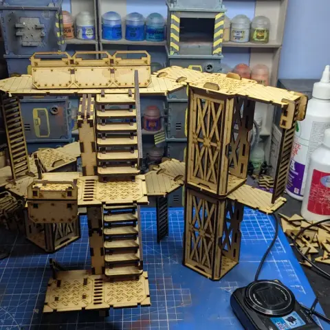A detailed assembly of modular terrain pieces made from laser-cut wood, featuring multiple levels, staircases, and platforms. The structure includes various textures and patterns, with a mix of open and enclosed areas, showcasing a futuristic industrial design. In the background, paint bottles and tools are visible, indicating a workspace for painting and customizing the terrain.