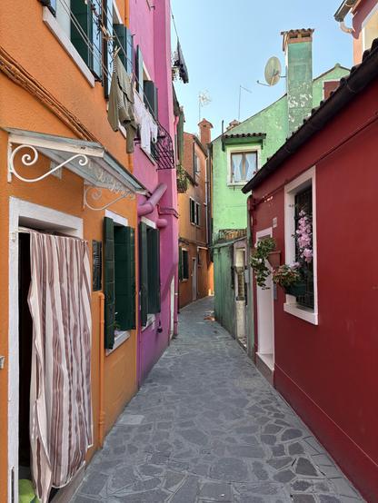 Brightly coloured houses in Burano on both sides of a narrow path. Houses are painted in variety of bright colours including orange, bright pink, maroon and pale green.
