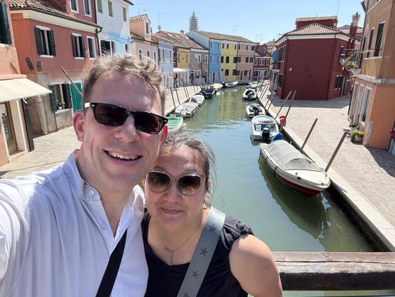 Picture of me and my wife standing on bridge crossing canal in Burano. The canal has many parked boats and brightly coloured houses are visible in background.