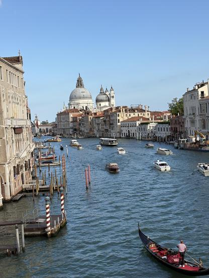 View of Grand Canal in Venice showing a mixture of boats and a gondola in foreground. Blue sky frames the top half of the photo.
