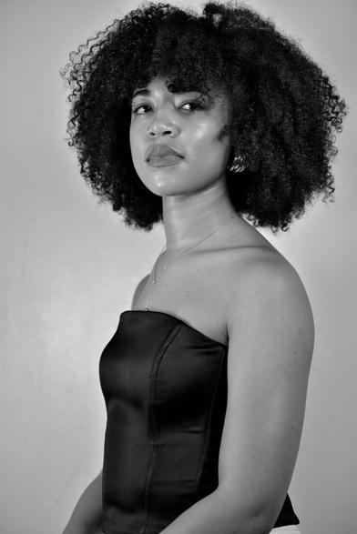 Waist-up portrait of a Black woman sitting down. She's angled facing the left of the camera as she stares off