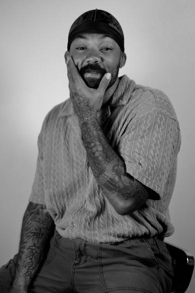 Waist-up portrait of a Black man sitting down. He's facing the camera with his left hand up to his face seemingly mid-conversation
