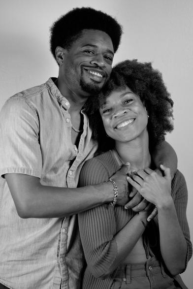 Waist-up portrait of a Black couple. The woman is sitting down and the man is standing up behind her and to the left. He has his arms wrapped around her shoulders while she leans towards his body.