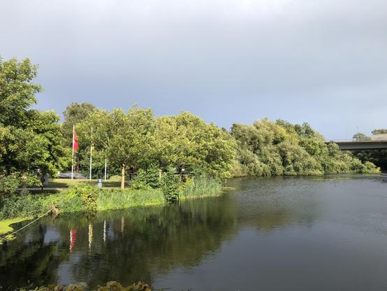 Düstere Wolken , dunkles Wasser. Sonne aber in den Bäumen und Sträuchern am Ufer.