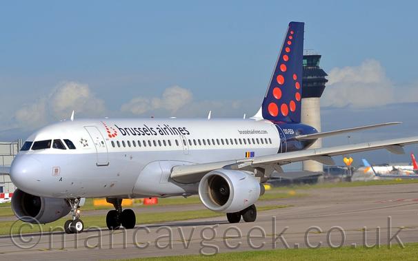 Side view of a twin engined jet airliner taxiing from right to left, turning onto a grey runway.
The plane is largely white, with a grey belly, and black Brussels Airlines titles on the upper forward fuselage, next to a series of red dots arranged to form a lower-case letter "b".
The rear fuselage and tail are dark blue, with more of those red dots forming 
a larger letter "b".
A black web address, "BrusselsAirlines.com"is on the upper rear fuselage, next to the white registration "OO-SSA".
A grey taxiway lined with green grass fills the foreground, leading off to a terminal building in the distance on the right, with several planes parked in front.
A tall grey tower with a heavily glazed, black top piece stretches into the sky.
Blue sky with large patches of white and grey cloud fill the rest of the frame.