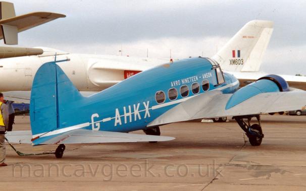 Side view of a twin propellor-engined post-World War 2-era airliner parked on a concrete apron, facing to the right and slightly away from the camera.
The plane is mostly blue, with a a thin white stripe running along the body, over-laid with the white registration "G-AHKX" on the rear fuselage, and white text "Avro Nineteen - Anson" on the upper forward fuselage.
The wings are an almost metallic grey, while the wing-mounted engine pods, which also house the retractable main landing gear, are the same blue as the fuselage, with the cowling around the engine being a darker blue.
The tail of a larger, sandy-brown coloured plane is visible on the left of the frame, while a large, white, 4 jet engined bomber aircraft is parked so if fills the rest of the background, with red engine covers over the engine inlets in the wing roots, and the black serial "XM603"under a red, white, and blue fin flash on the tail.
Grey sky completes the rest of the frame.