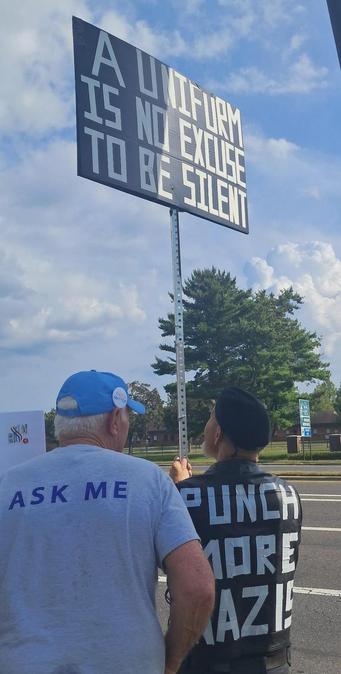 Two men are standing beside a roadway.  One has a tee shirt with the words, "Ask Me," on the back.  The other man is wearing a shirt with the words, "Punch More Nazis," on the back.  He is holding high a sign on a pole that reads, "A Uniform Is No Excuse To Be Silent."