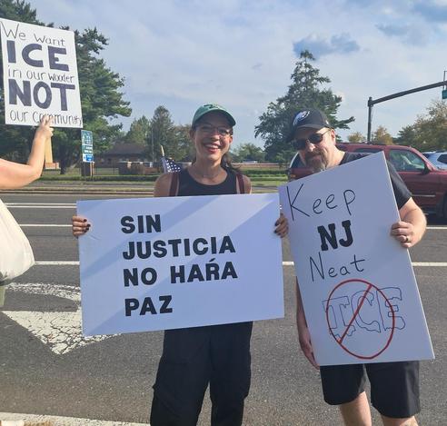 A woman and a man are holding protest signs as they stand along a roadway.  The woman's sign reads in Spanish, "Sin Justicia, No Hara Paz," which means in English, "No Justice, No Peace." The man's sign reads, "Keep NJ Neat."  The word ICE is encircled in red with a red line crossing it out.  Another person, who is only partially seen, is holding a sign that reads, "We Want Ice In Our Wooder, Not In Our Community." (Wooder is a funny way some New Jerseyans say "water").