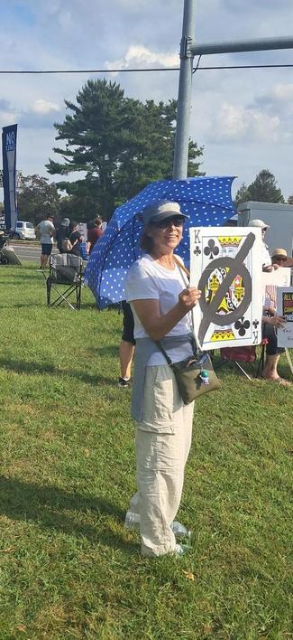 A woman is holding a protest sign that looks like a King of Clubs playing card.  A circle with a line through it is drawn over it, meaning No Kings!
There are many more protesters gathered with her.