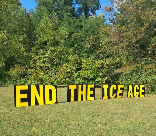A huge sign is displayed on the grass.  A forest is behind it.  The words are in yellow capital letters with black trim.  The sign says, "END THE ICE AGE."