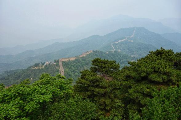 A high-angle long shot shows the Great Wall of China winding its way across a lush, mountainous landscape. The wall, constructed from grey stone, snakes along the ridges and valleys of rolling hills that are covered in a dense mix of deciduous and coniferous trees. The sky is hazy and overcast, casting a soft, muted light over the scene. In the foreground, the tops of green trees fill the lower portion of the frame. In the distance, the mountains fade into the mist, their outlines becoming less distinct.