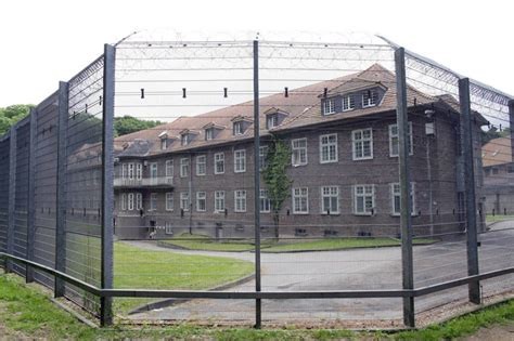 Vierstöckiger altes Gebäude mit Giebeln. Rot-braune Ziegel, davor ein mehrere Meter hoher Zaun mit Stacheldraht auf dessen Krone. Zwischen Zaun und Gebäude ein bisschen grüner Rasen und ein planierter Spazierweg
