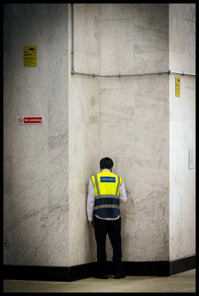 Security guard in yellow and navy high-visibility vest with "SECURITY" text standing in corner of white marble-walled corridor or building interior, with yellow CCTV warning signs and "No smoking" sign visible on walls in London.