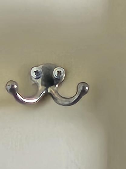A silver coat rack with 2 extended hooks ending in round balls is attached to a darkened off-white wall with 2 Phillips head screws.