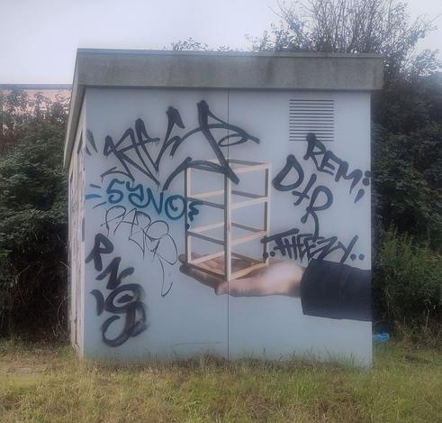 Photo of the side of a small shack โ perhaps a transformer station โ with a mural. A hand is painted on a light grey background, palm facing upwards, wrist/forearm covered by the sleeve of a jacket. On the palm is a geometric shape, resembling an open wooden mini bookcase with three shelves.
Several graffiti tags have been written over the mural, mostly in black.