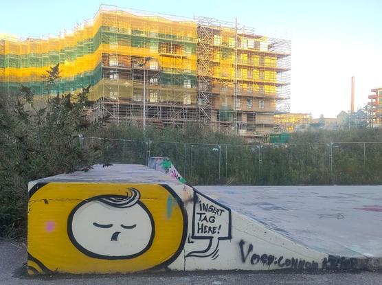 A photograph of graffiti artwork on the side of a low quarter pipe/edge of a skate park. It is only the head of a ‘Friendly Hero’ character, consisting of a white circle with closed eyes, open mouth, and a tuft of hair standing up, surrounded by a yellow band like a hood. To the right of the head is a speech bubble with ‘insert tag here’ written inside.

To the right, you can see the concrete floor of the skate park. Behind it and to the left are bushes, and in the background, a multi-storey building under construction, covered in scaffolding and surrounded by a green and yellow striped net. The yellow colour looks particularly bright in the sunlight.