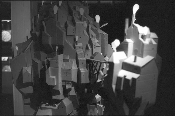 A black and white photograph of a miniature city made out of cardboard. Small windows and artificial smoke made out of paper gives the illusion of scale.