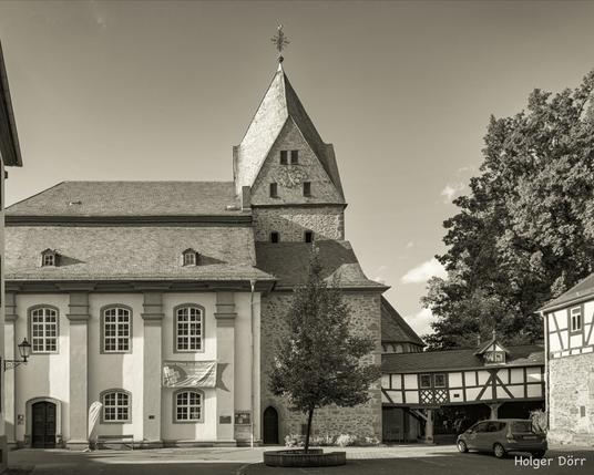 Die Evangelische Stadtkirche in Laubach erhebt sich mit ihrem wuchtigen Turm und den barocken Fenstern über den Platz der Altstadt. Im Vordergrund ein Baum und Fachwerkdetails, die der Szene eine einladende, historische Atmosphäre verleihen. Das Schwarz-Weiß des Fotos unterstreicht die Zeitlosigkeit und den Charakter des Bauwerks.