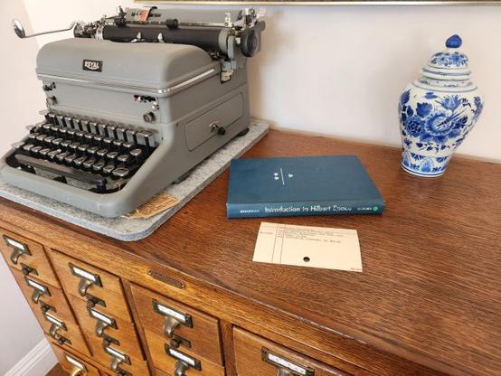 A blue textbook with three crowns on the cover and a faded spine that reads Introduction to Hilbert Spaces sits on an oak library card catalog between a Royal KMG typewriter and a blue vase.