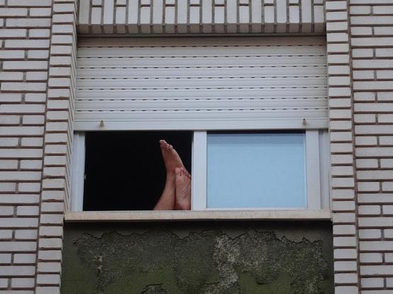 Plano cerrado de una ventana abierta, con la persiana a medio bajar, en la fachada de un edificio (ladrillos blancos). Apoyados en el alféizar y el marco se ven los pies de una persona.