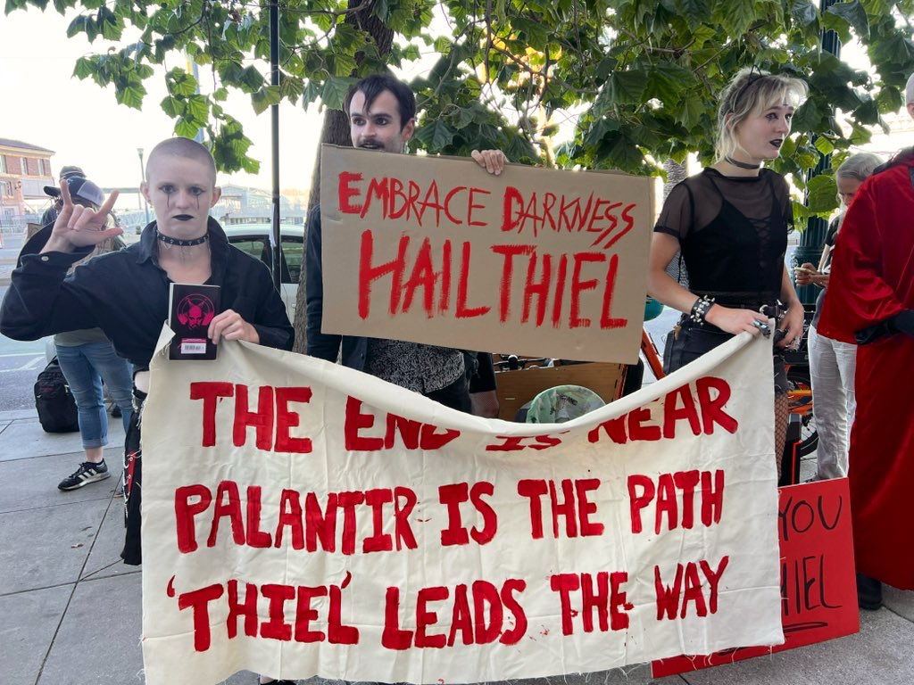 Costume Satanists holding signs. “Embrace darkness, hail Thiel.” “Palantir is the Path.”