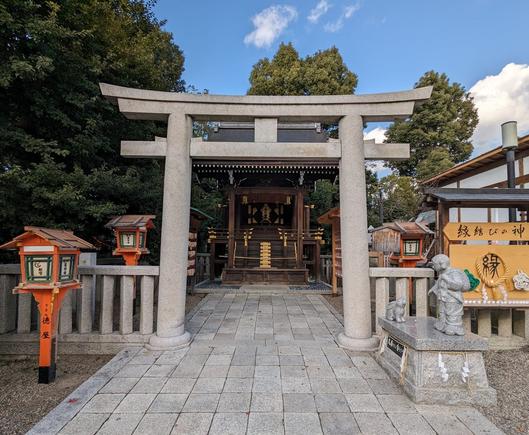 A stone torii leading to a small shrine. Red lantern to the left. In the right a stone statue of the kami Okuninushi holding a sack over his shoulder and looking down at a rabbit.
