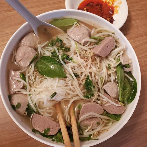 A bowl of Vietnamese pho, a clear broth with rice noodles, slices of meat, bean sprouts, herbs, and meatballs, served with a small dish of chili oil and chopsticks, on a wooden table.