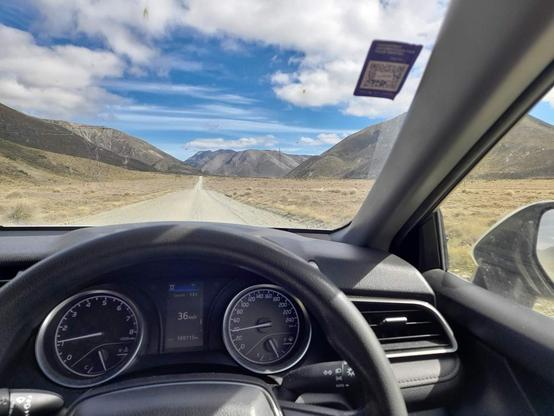 L’image invite au voyage, capturant un instant sur une route solitaire qui s’étire droit vers l’horizon, bordée de montagnes silencieuses et tapissées d’herbes dorées par le soleil. Par le pare-brise, le ciel néo-zélandais dévoile ses nuages vaporeux et ses éclats d’azur, comme une toile offerte à l’aventure.
À l’intérieur, l’habitacle est baigné de lumière, le volant posé devant ce panorama sauvage, promesse de nouveaux départs et de liberté. Sur cette piste poussiéreuse, la voiture semble flotter entre deux mondes : celui du confort moderne et celui de la nature indomptée, invitant l’esprit à s’évader dans la tranquillité immense des paysages de Nouvelle-Zélande.