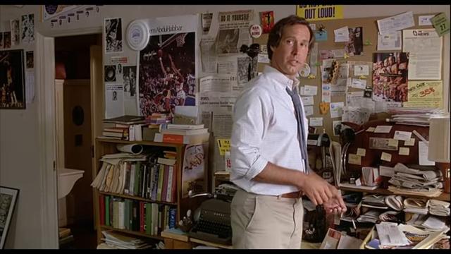 Standing, Fletch looks up from his desktop. Behind him in the middle of his home desk sits a Royal HH typewriter.