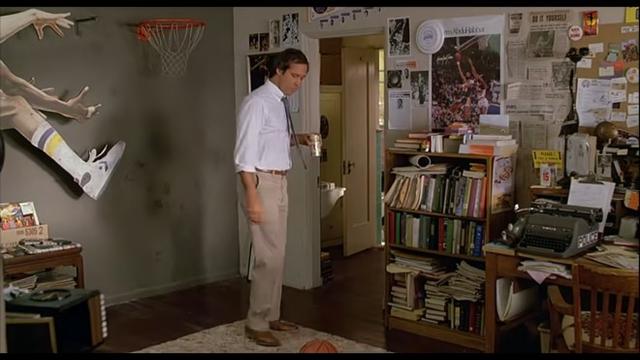 Fletch with a beer in hand eyes a basketball on the floor. In front of him on the desk is a brown Royal HH standard typewriter.