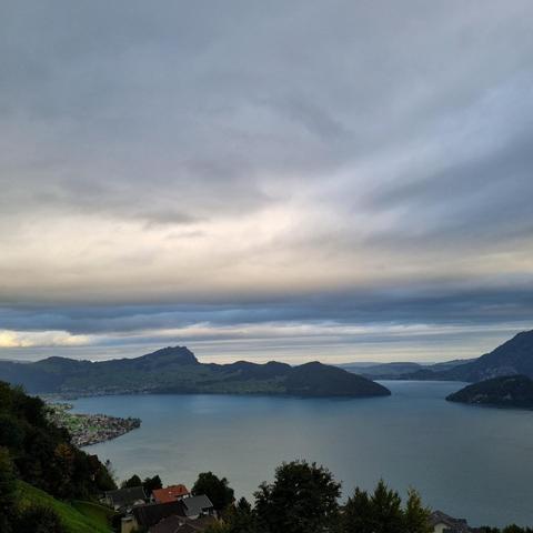 View of a lake, the shores are visible as silhouettes. Above it is a dense cloud cover, the middle layer of which glows brightly.