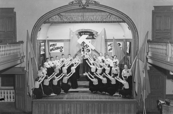 A black and white image of a stage with young people, wearing white and with a black hammer on their chest, doing some form of choir choreography around a large white hammer.