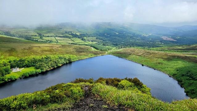 A wide-angle aerial shot from a misty mountain top overlooking a tranquil, dark-blue lake. A small white house with a grey roof is nestled in a grove of trees on the left shore. The surrounding hills are a patchwork of green fields, brown heath, and dense woodlands, extending into the distant mountains, where a low-lying mist obscures the peaks.